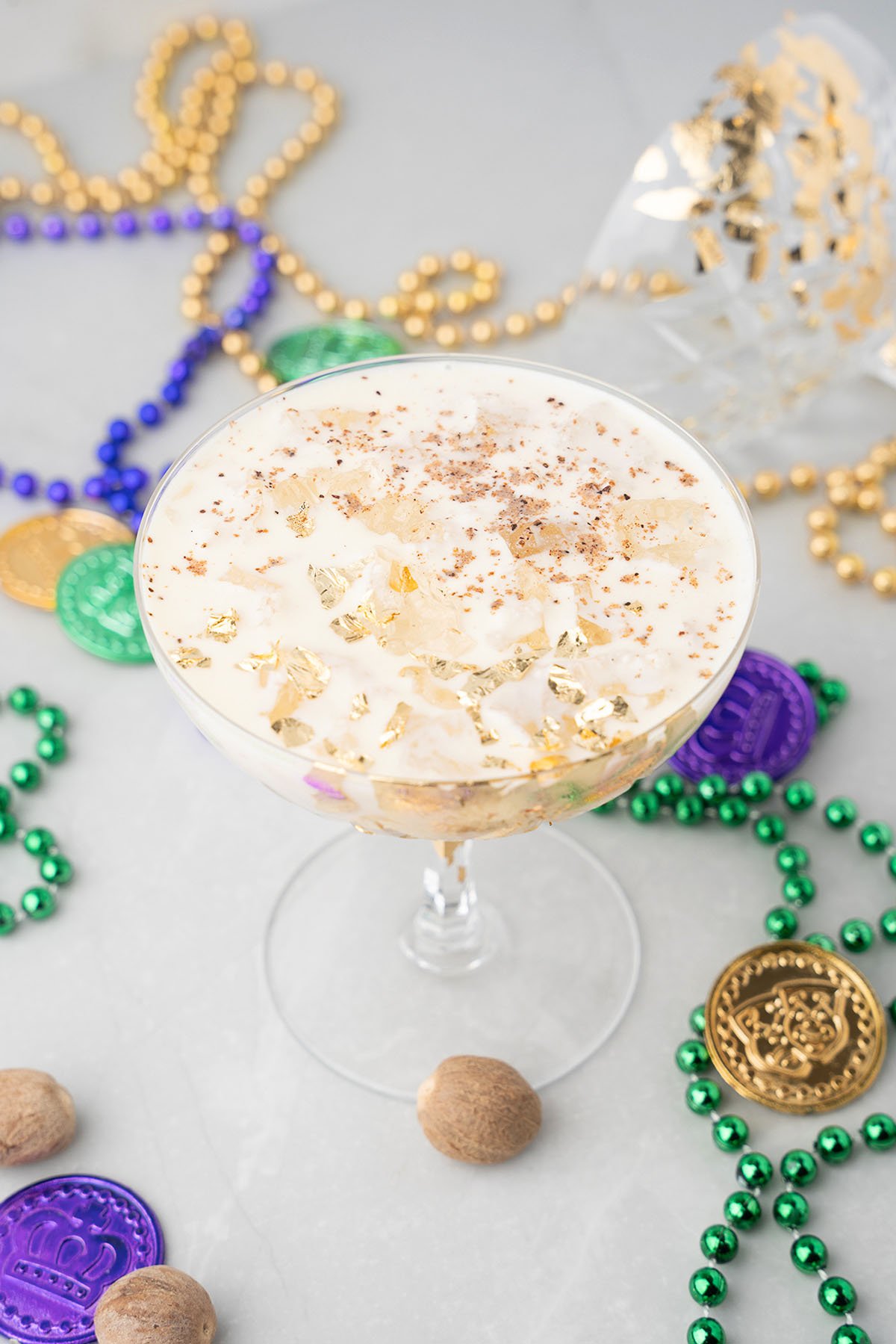 A glass of creamy New Orleans milk punch topped with gold flakes, surrounded by Mardi Gras beads and coins.