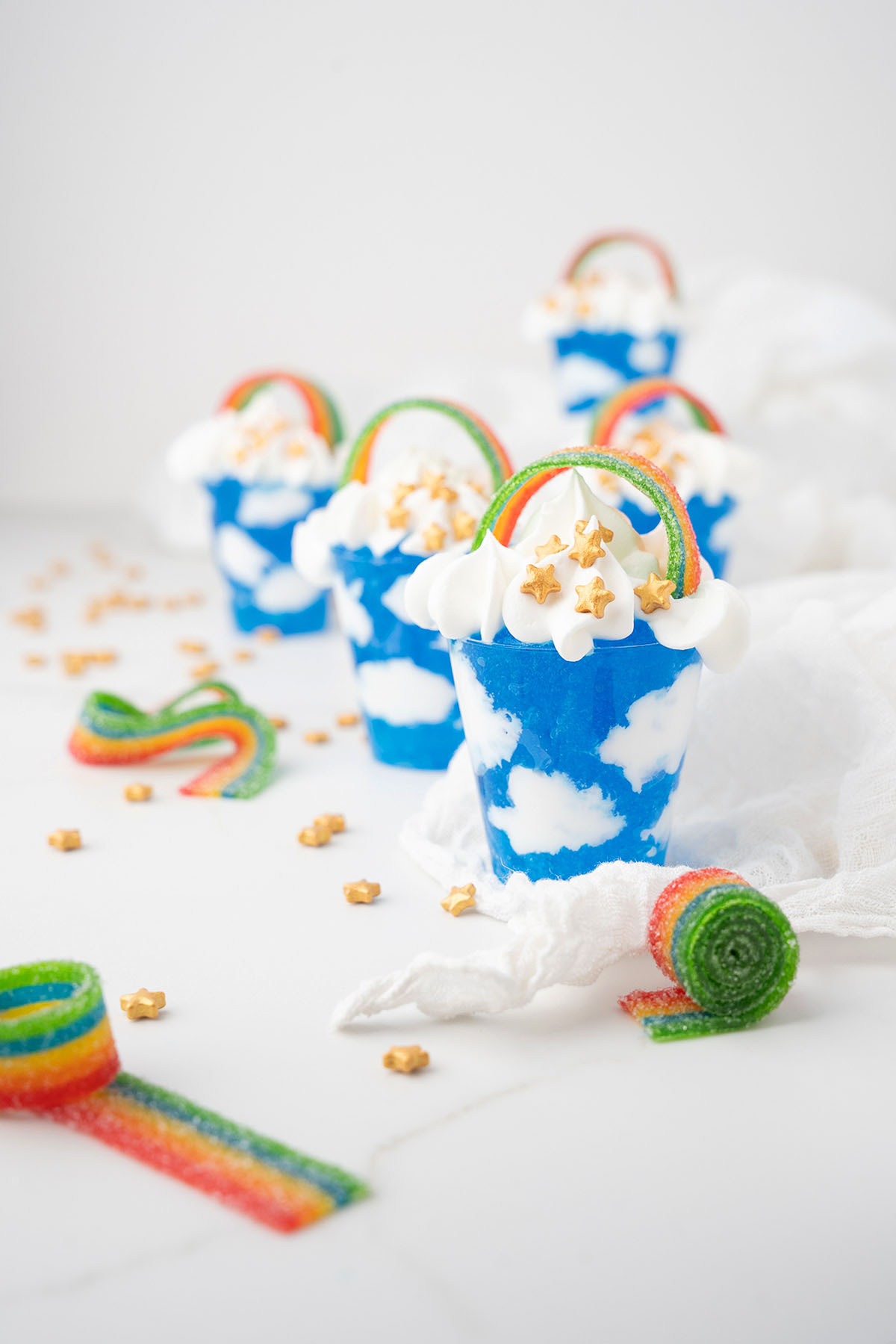 Blue cupcakes with white cloud frosting, rainbow candy, and gold star sprinkles on a white surface&mdash;perfect for St. Patrick&rsquo;s Day or Pride Day celebrations, inspired by jello sky and cool whip clouds.