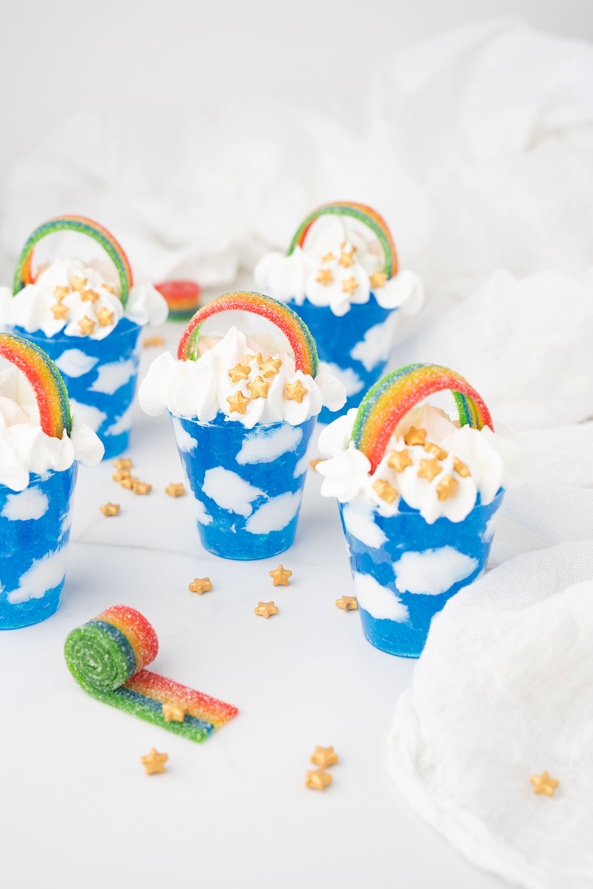 Blue cloud-themed desserts in cups, featuring Jello sky and cool whip clouds, are topped with star sprinkles and rainbow candy strips&mdash;perfect for Pride Day or a whimsical Rainbow Pot O' Gold Jello Vodka Shot treat.