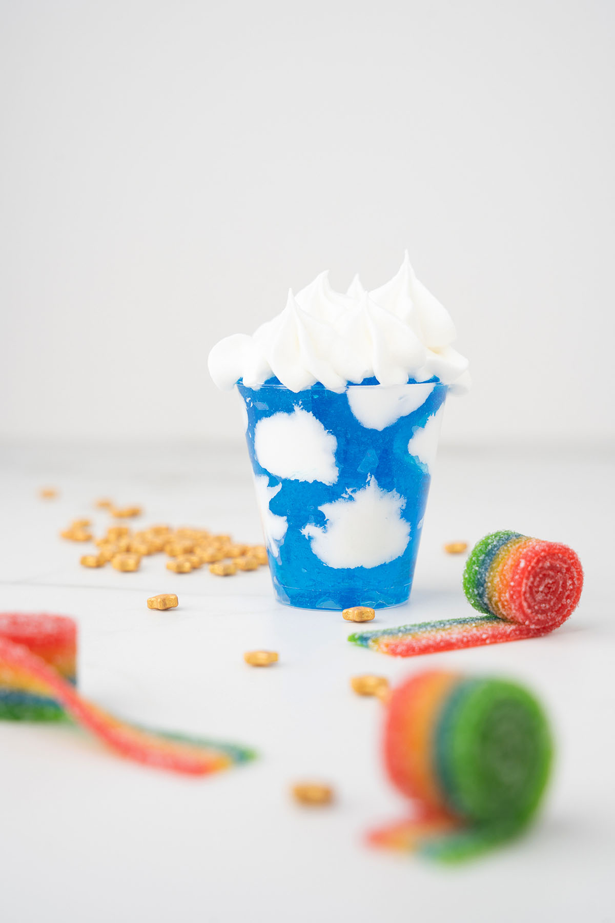A blue dessert cup with white cloud shapes, topped with whipped cream and surrounded by rainbow candy strips&mdash;perfect for Pride Day celebrations. Enjoy this festive treat inspired by Rainbow shots and Jello sky and cool whip clouds!.