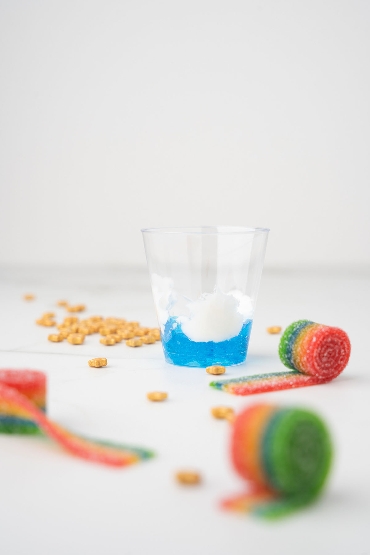 A clear cup with Jello sky and cool whip clouds, surrounded by rainbow candy rolls and scattered cereal.