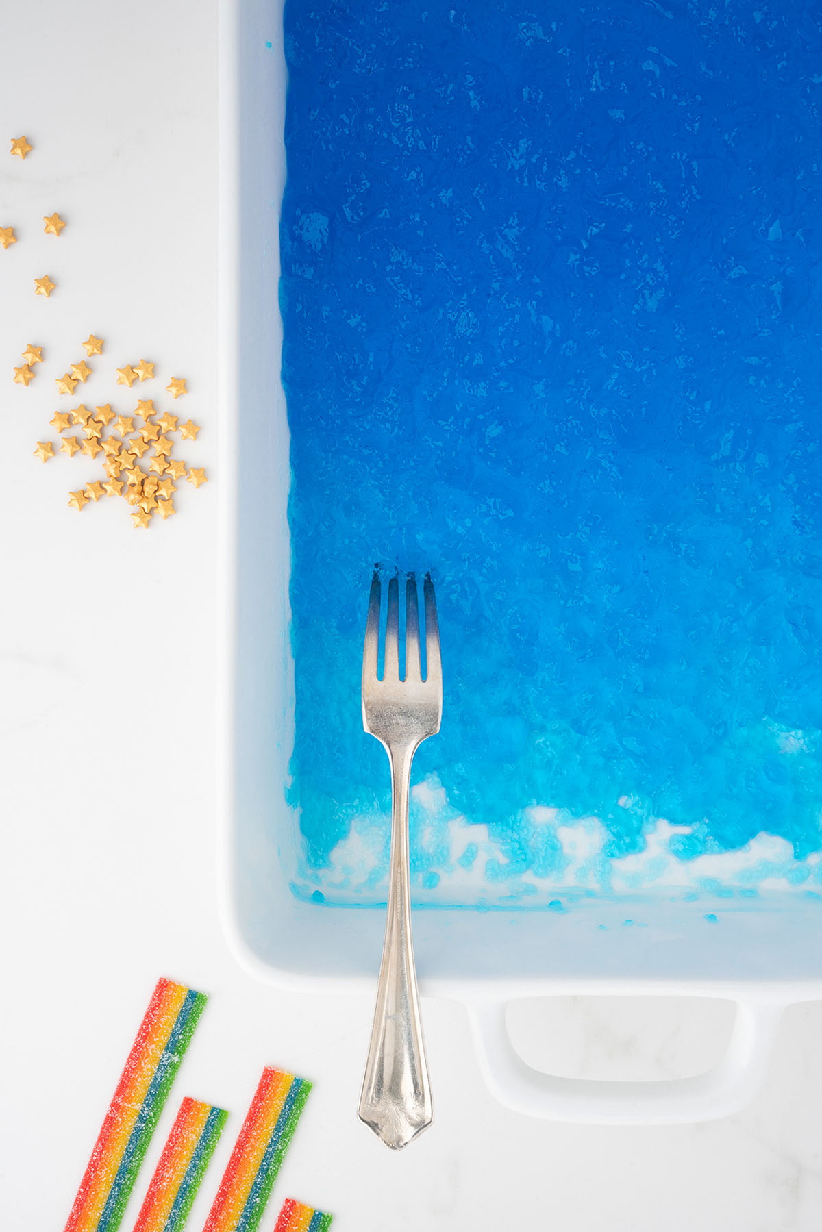 A fork in blue gelatin with star-shaped sprinkles and rainbow candy strips on a white surface&mdash;perfect inspiration for Rainbow Pot O' Gold Jello Vodka Shots at your next St. Patrick&rsquo;s Day or Pride Day celebration.