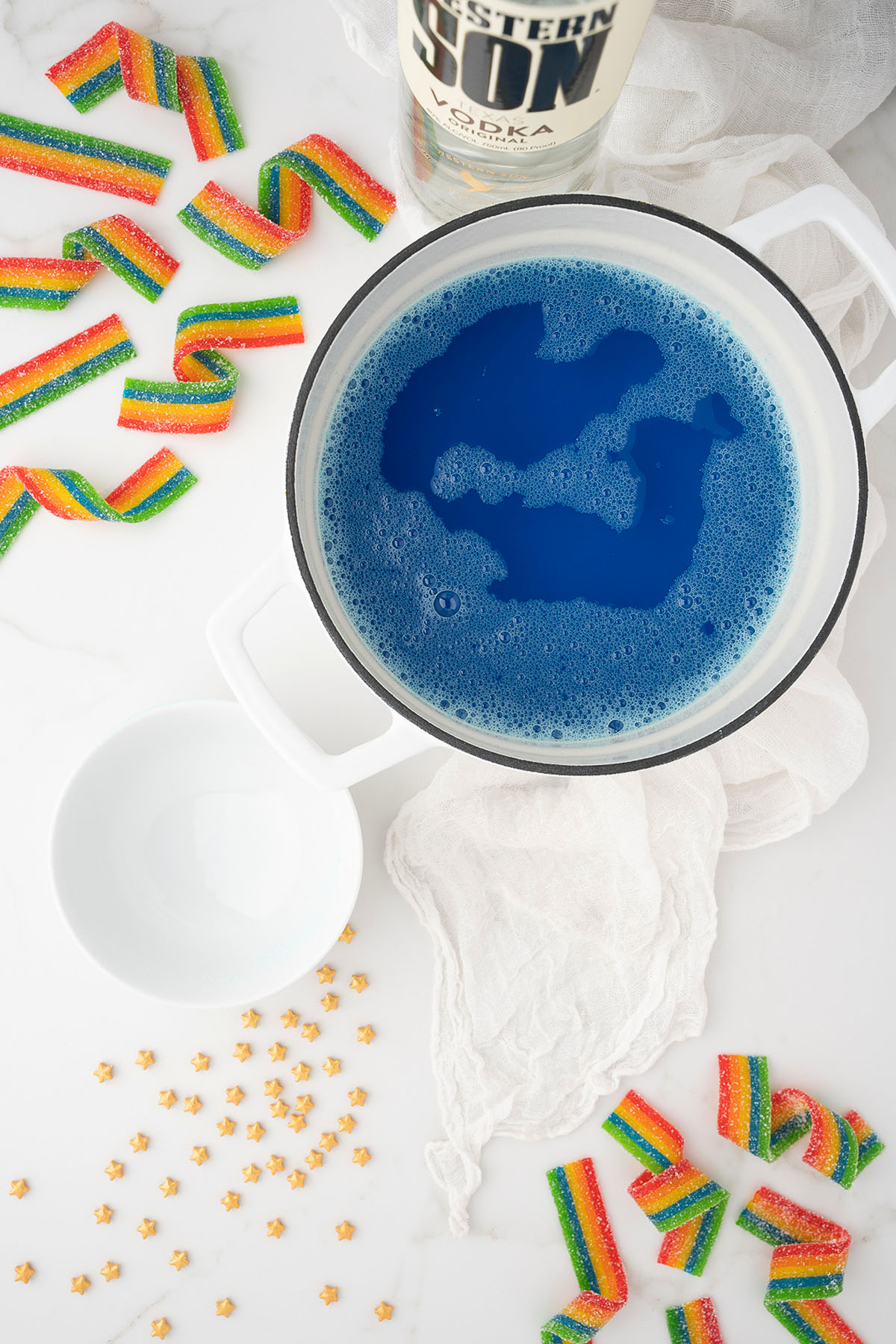 A white pot of blue liquid, rainbow candy strips, and gold star sprinkles on a white surface&mdash;perfect inspiration for festive Rainbow shots for St. Patrick&rsquo;s Day or Pride Day celebrations.