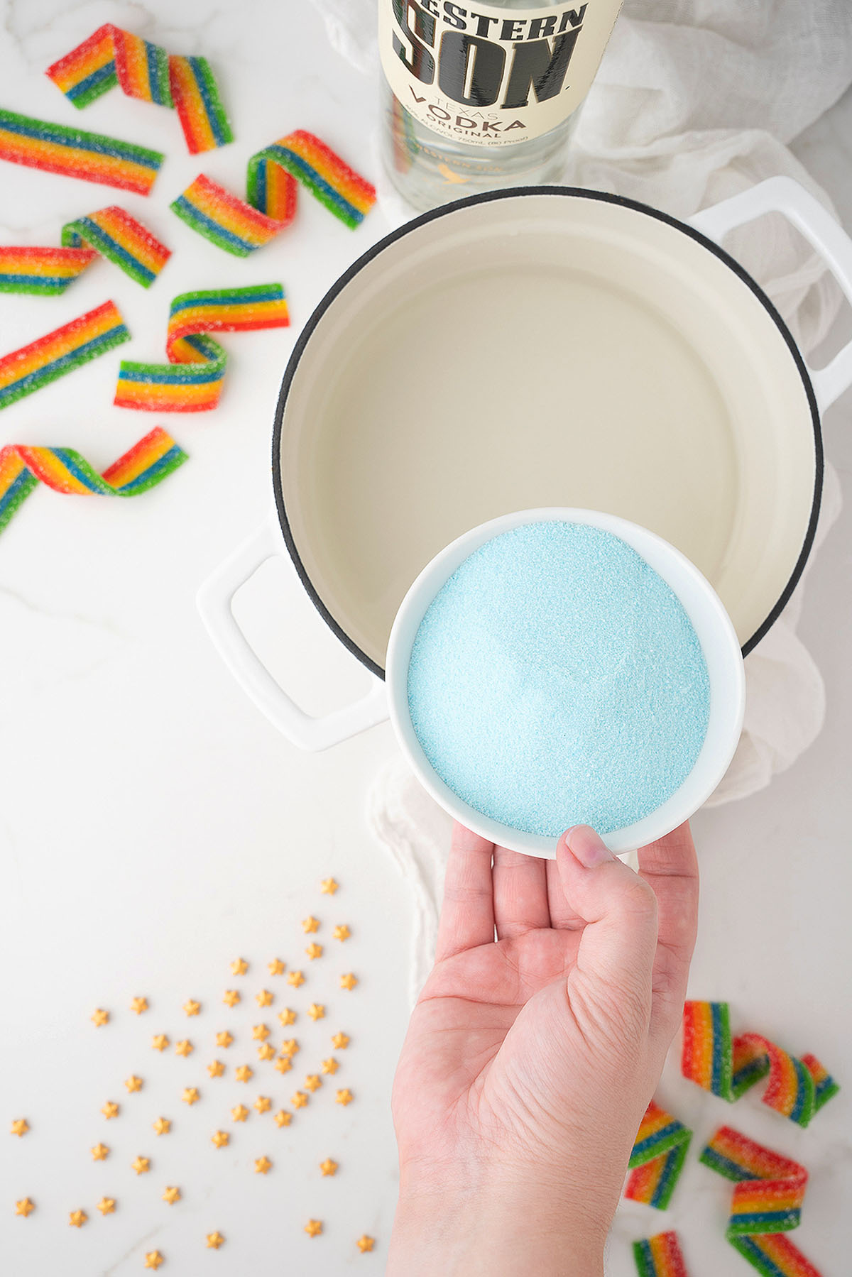A hand holds a bowl of blue powder over a pot of clear liquid, with rainbow candy and star sprinkles nearby&mdash;perfect for making Rainbow Pot O' Gold Jello Vodka Shots for St. Patrick's Day or Pride Day celebrations.
