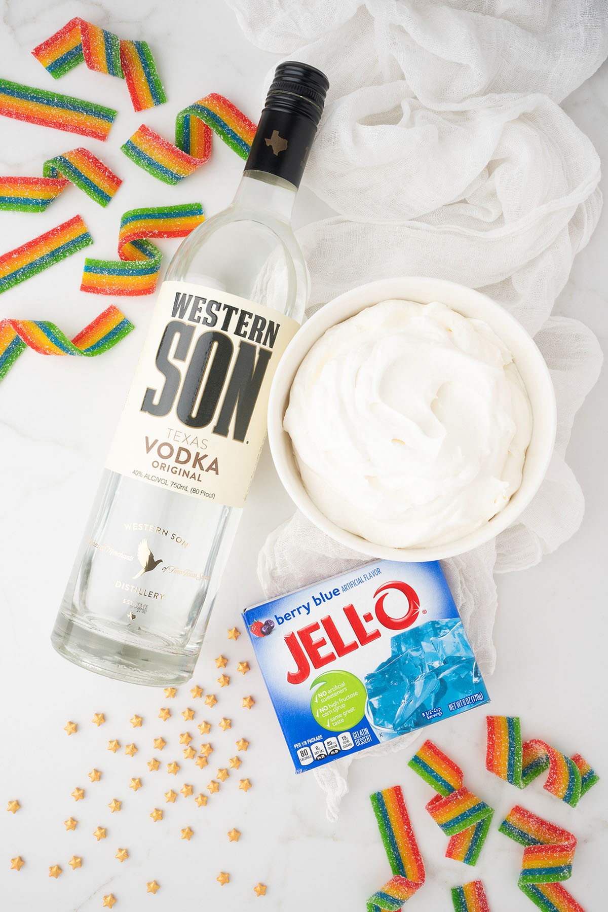 A bottle of vodka, a bowl of whipped topping, blue Jell-O mix, and rainbow candy strips on a white surface&mdash;perfect for making Rainbow Pot O' Gold Jello Vodka Shots for Pride Day.