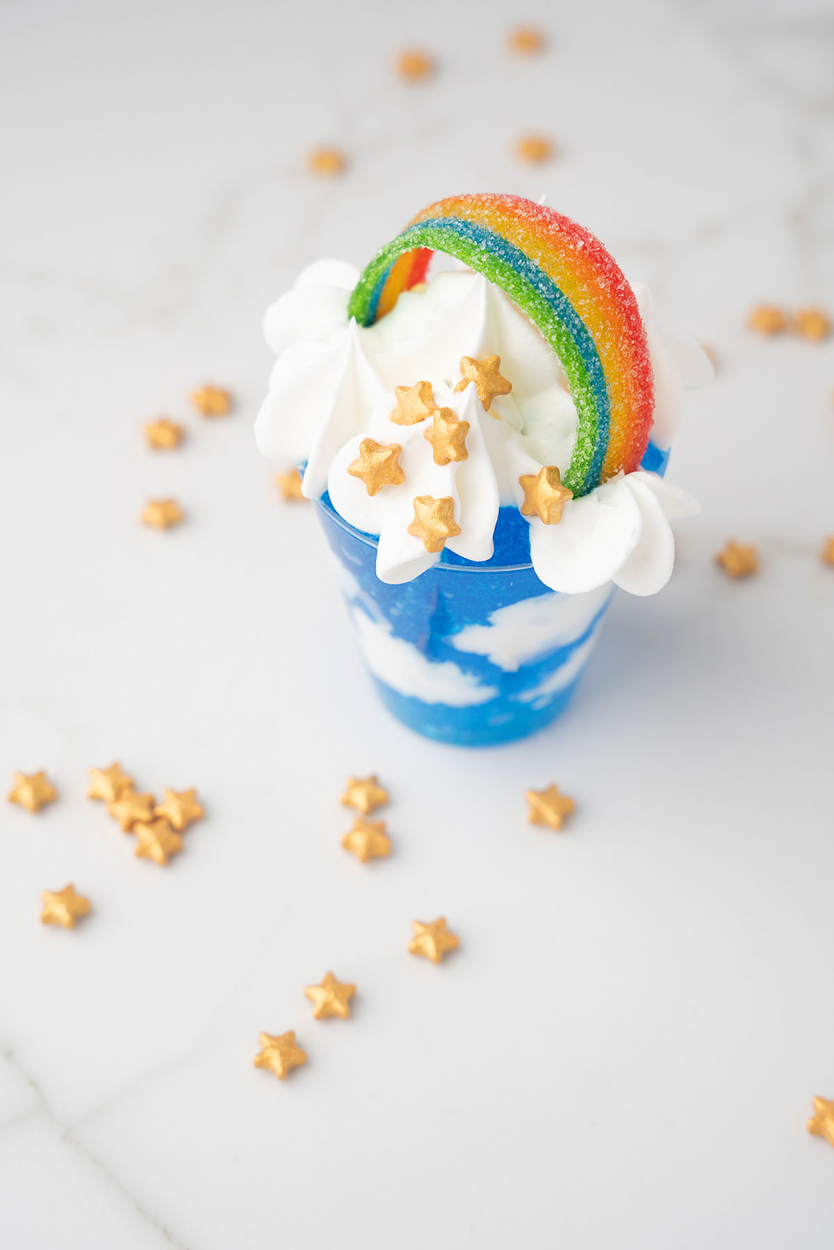 Blue dessert cup with whipped cream, a rainbow candy, and gold star sprinkles on a white surface&mdash;perfect for Pride Day or as festive Rainbow Pot O' Gold Jello Vodka Shots.