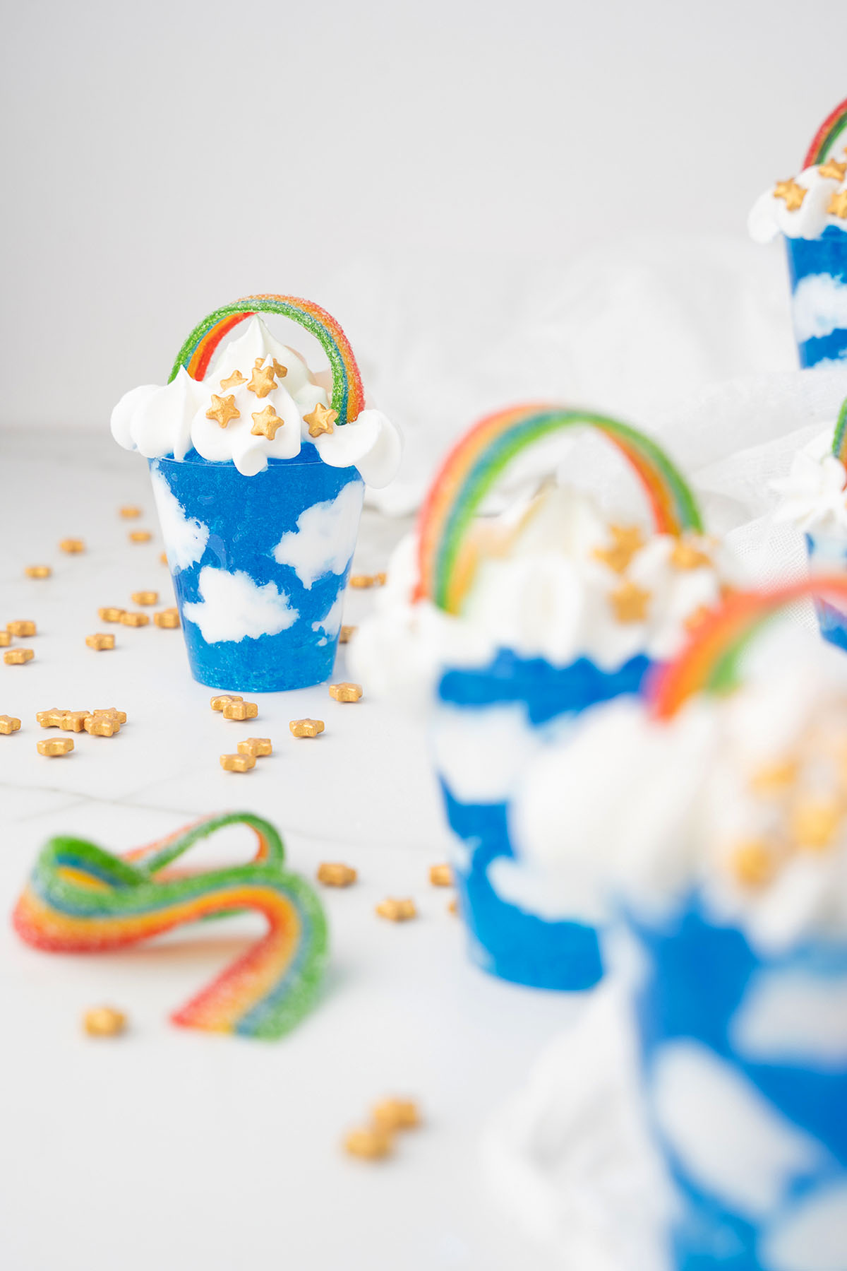 Blue and white cloud-themed dessert cups topped with whipped cream, candy rainbows, and star-shaped sprinkles&mdash;perfect for serving rainbow shots at your next St. Patrick's Day or Pride Day celebration.
