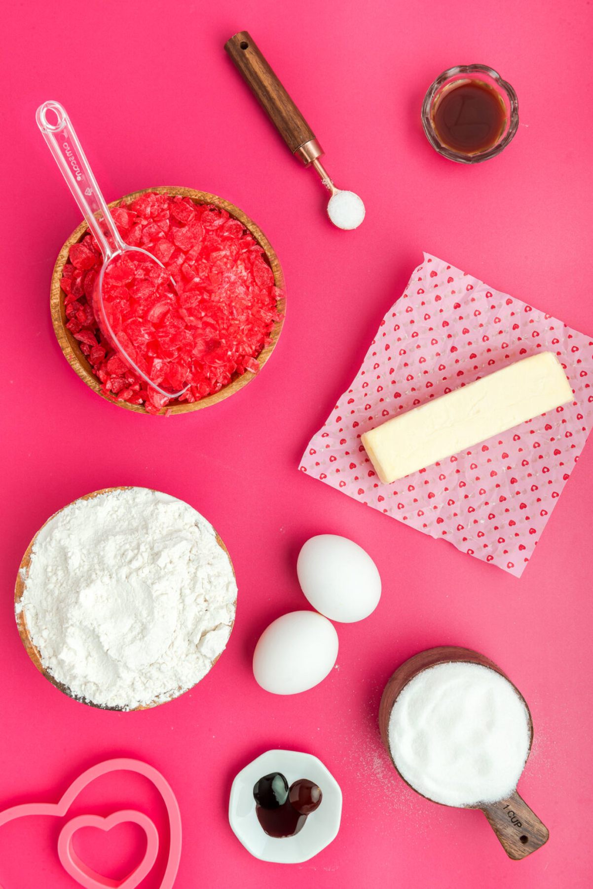 Baking ingredients on a pink background: flour, sugar, butter, eggs, red crumbs, vanilla, and heart-shaped cutters—perfect for making heart shaped sugar cookies or Stained Glass Valentine Heart Cookies with charming candy windows.