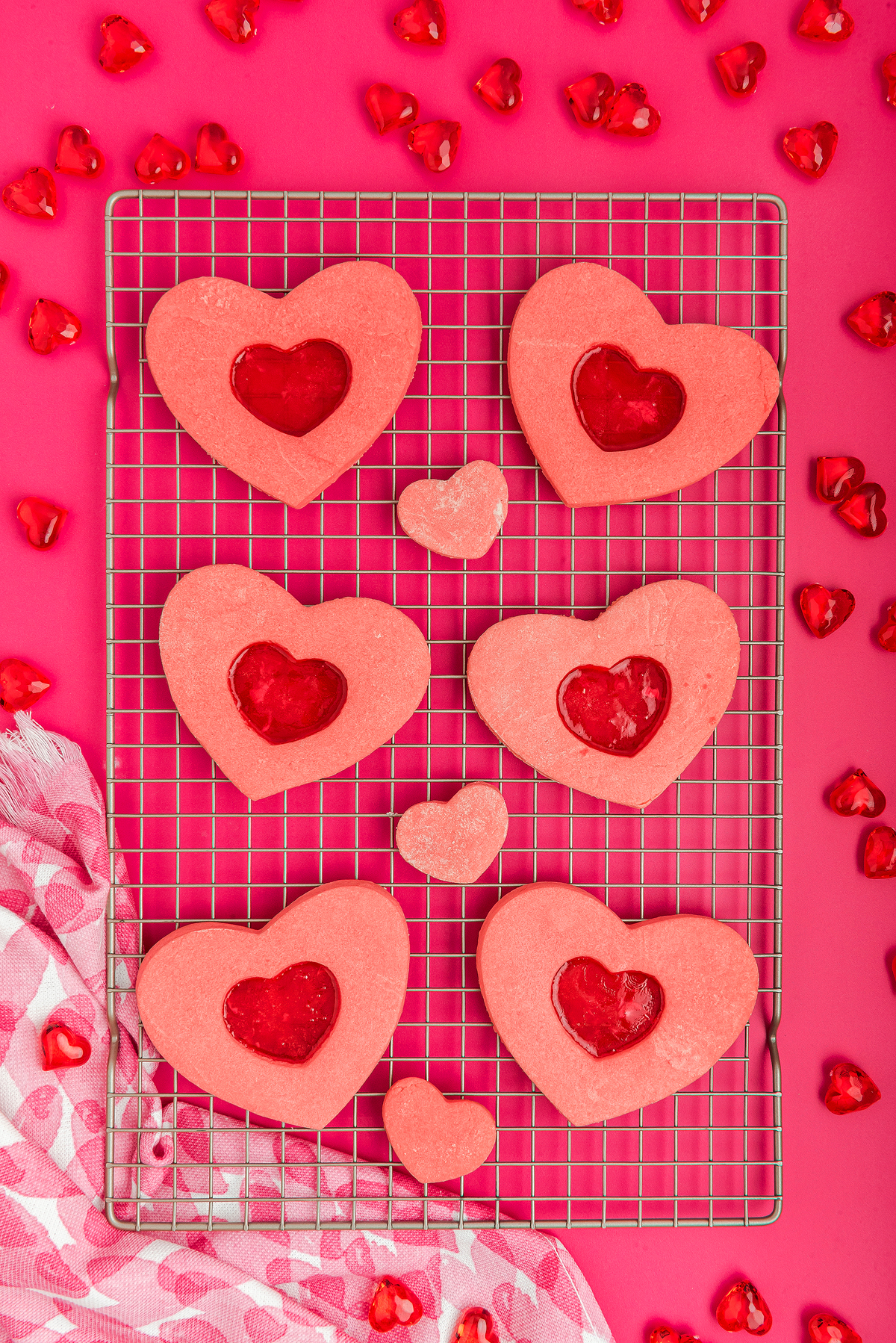 Stained Glass Valentine Heart Cookies: Pink heart-shaped cookies with red centers, made with Jolly Ranchers or other hard candy, rest on a cooling rack surrounded by small heart candies on a pink background.