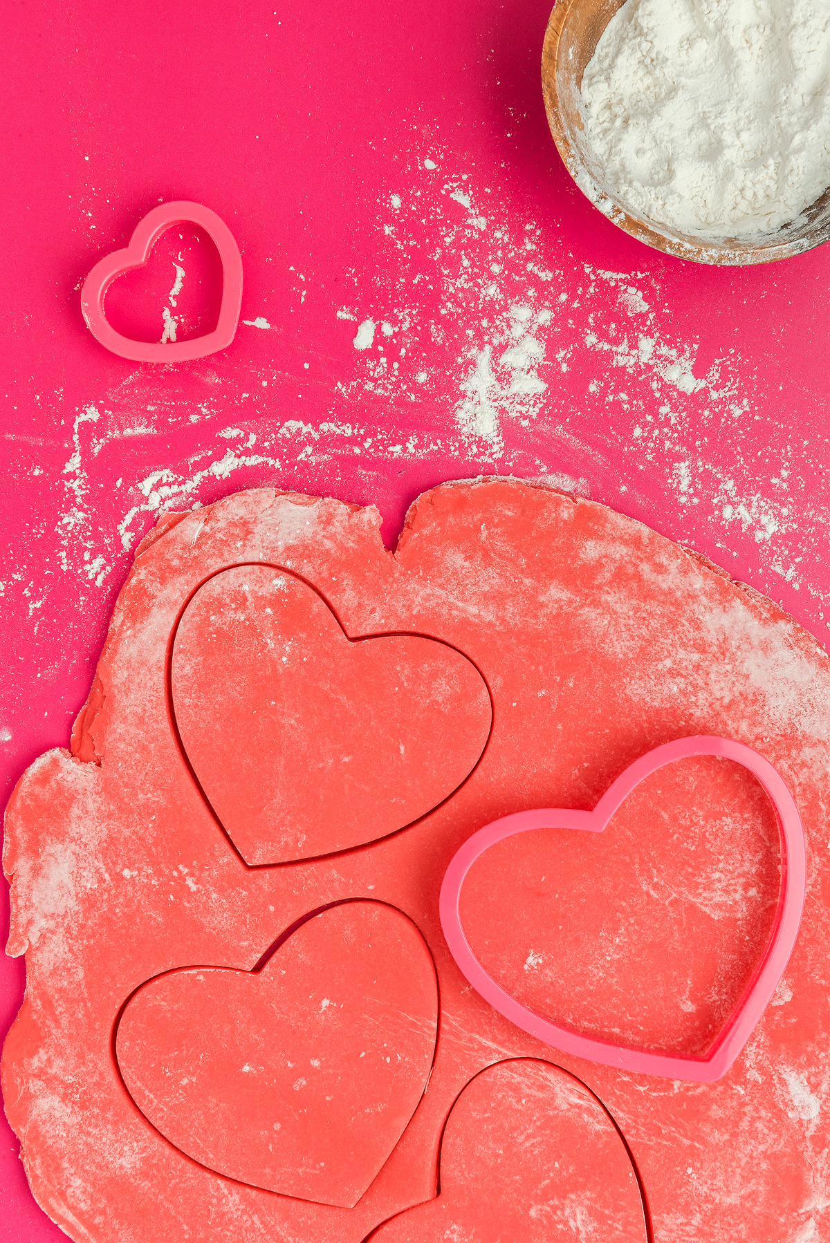Heart-shaped cookie cutters and dough on a pink surface with a bowl of flour in the corner, perfect for making Stained Glass Valentine Heart Cookies or heart shaped sugar cookies with candy windows.