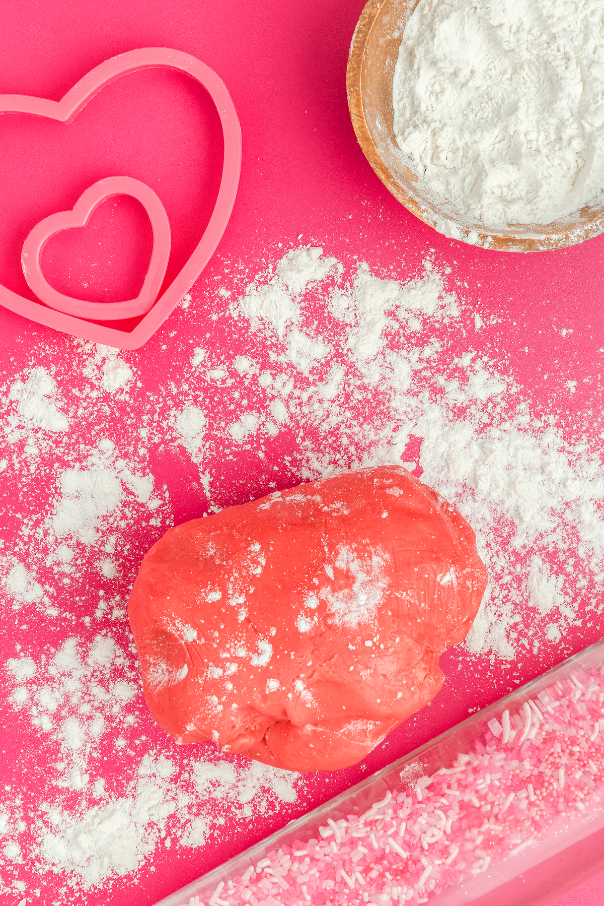 Red dough ball on pink surface with heart-shaped cutters, flour, and sprinkles scattered around—perfect for making stained glass Valentine heart cookies with colorful candy windows.