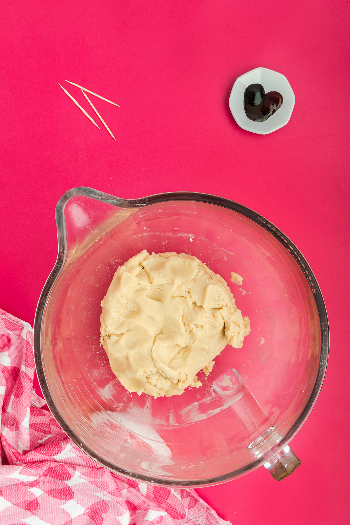A glass bowl with cookie dough on a pink surface, beside toothpicks, red food coloring, and a checkered towel—perfect for making Stained Glass Valentine Heart Cookies featuring candy windows made with Jolly Ranchers or other hard candy.