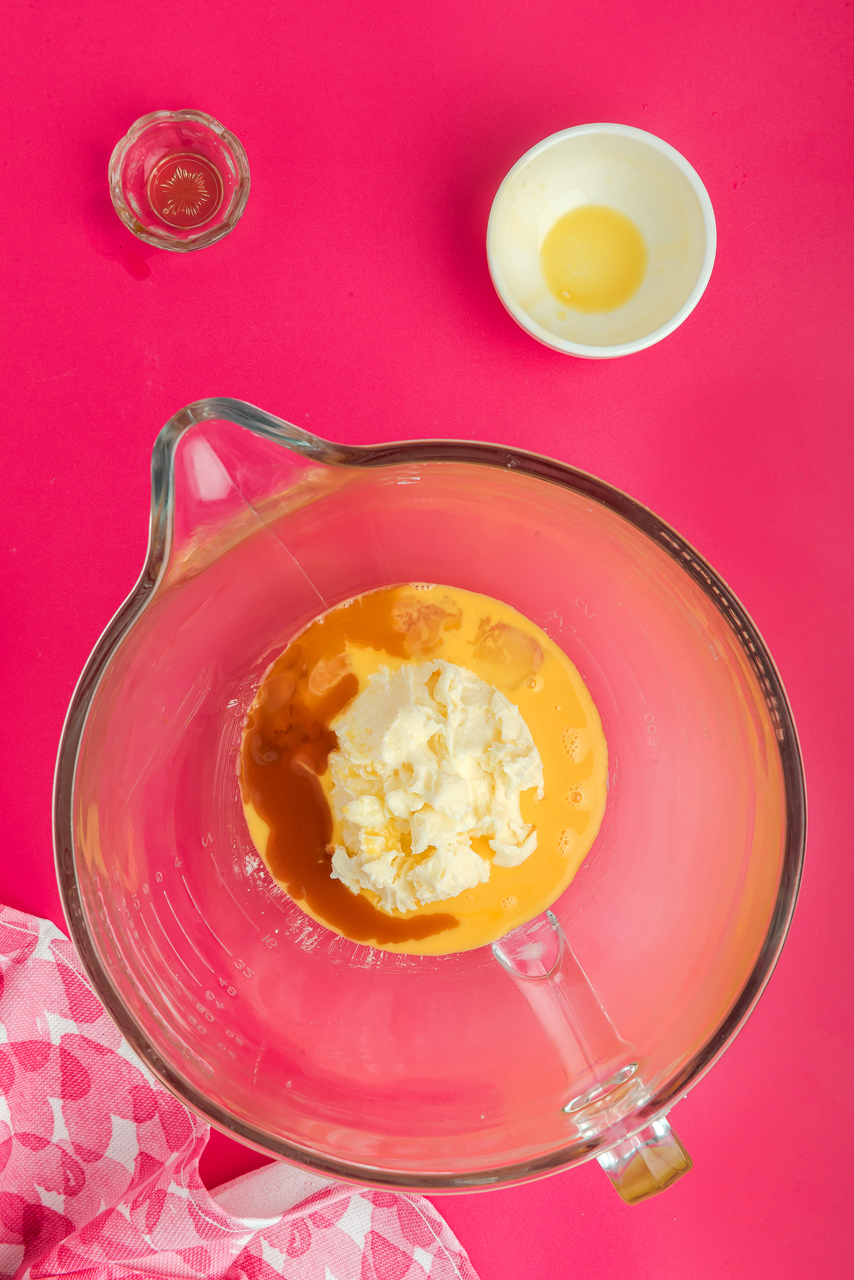 A glass bowl with eggs and cream cheese on a pink surface, with small bowls of vanilla and melted butter nearby—perfect for making Stained Glass Valentine Heart Cookies made with Jolly Ranchers or other hard candy.
