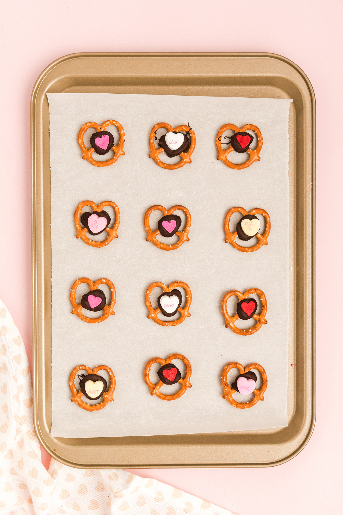 A baking tray with 12 heart-shaped Valentine Pretzel Snack Bites, each topped with chocolate and colorful candy hearts—perfect as a sweet Valentine’s Day treat.