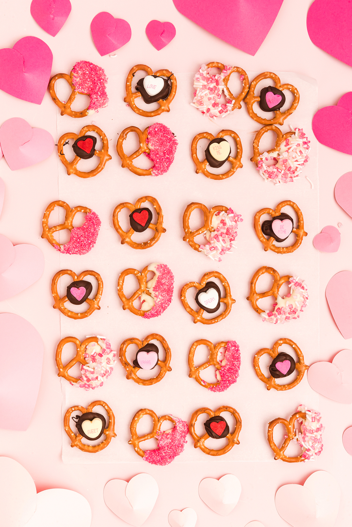 Heart-shaped Valentine pretzels with pink, red, and white decorations on a pink background with paper hearts make the perfect Valentine’s Day treat.