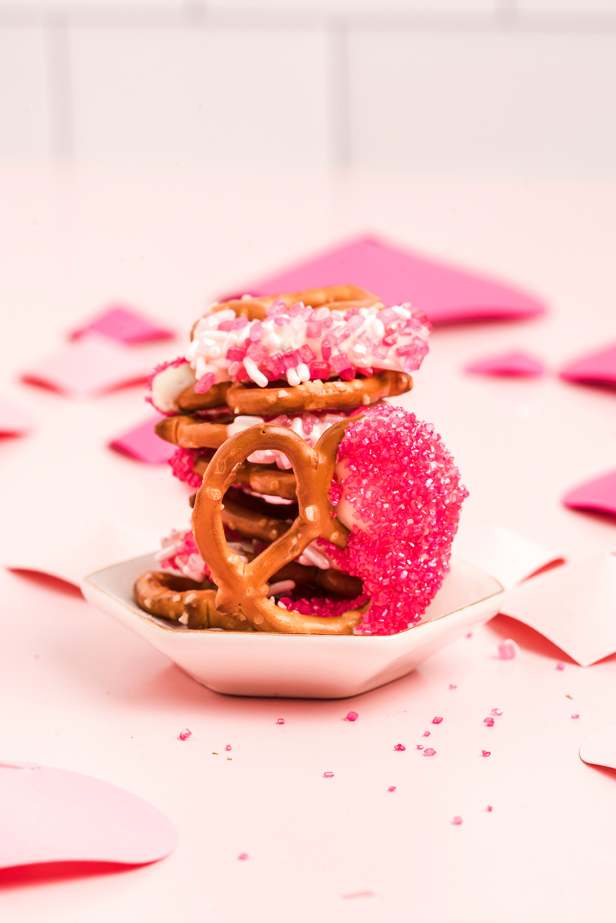 A stack of Valentine pretzels decorated with pink, white, and red sprinkles sits on a small white dish, making the perfect Valentine’s Day treat.