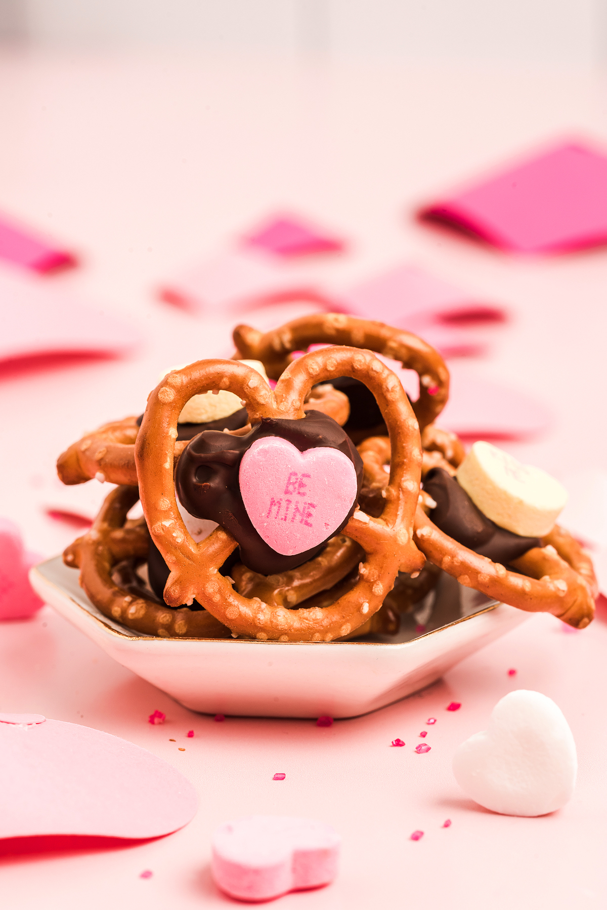 Heart-shaped Valentine pretzel snack bites with chocolate and pink candy hearts on a small dish, surrounded by festive heart decorations—an adorable Valentine’s Day treat.