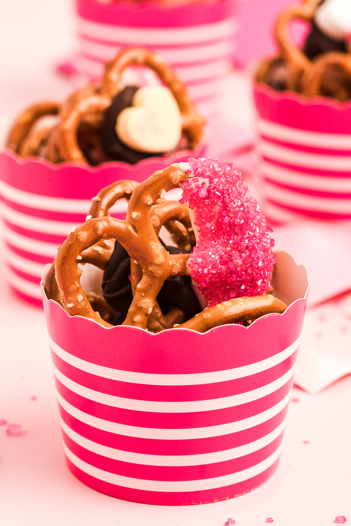Close-up of Valentine pretzels and chocolates in a pink-striped cup, topped with a pink sugar-coated treat—perfect as a Valentine’s Day treat.