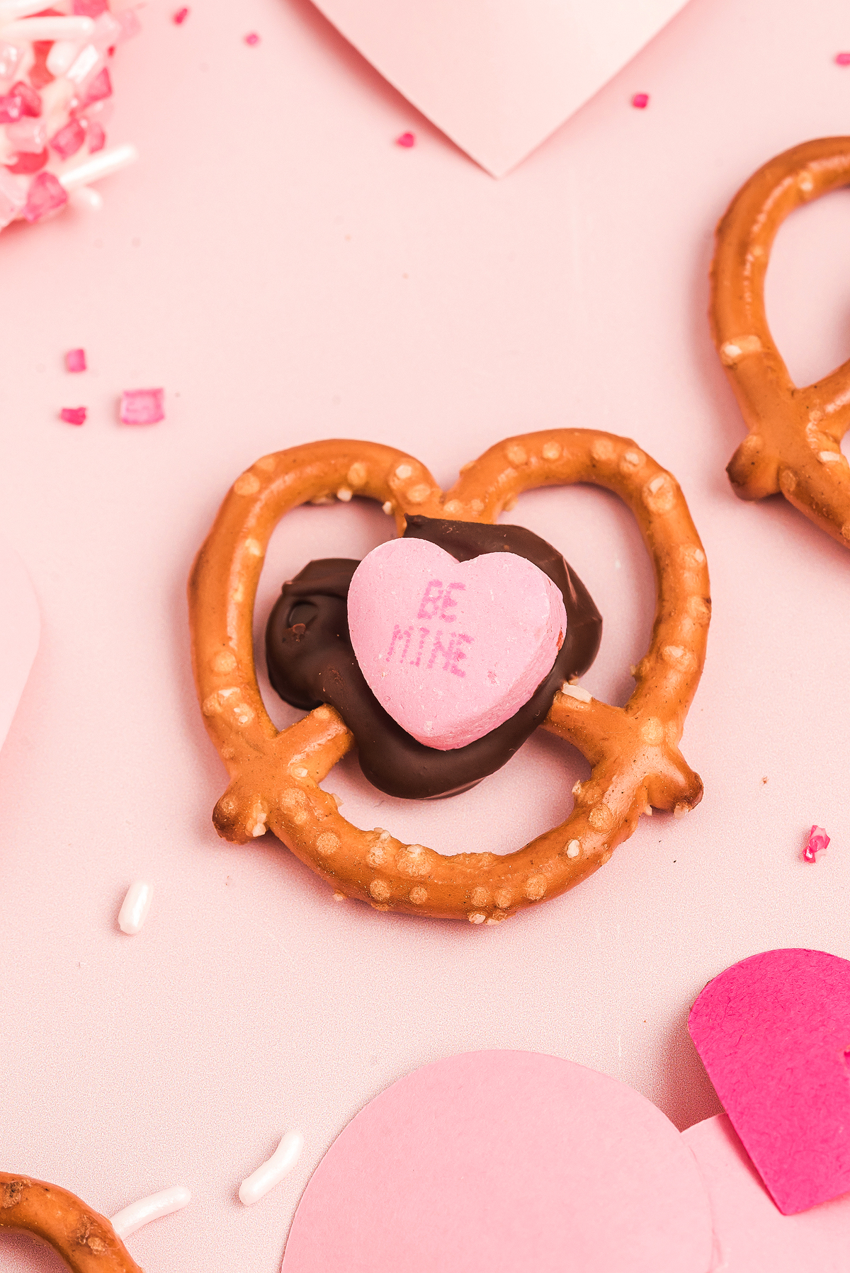 A Valentine Pretzel Snack Bite topped with chocolate and a pink candy heart that says "BE MINE" sits on a pink background—a perfect Valentine’s Day treat.