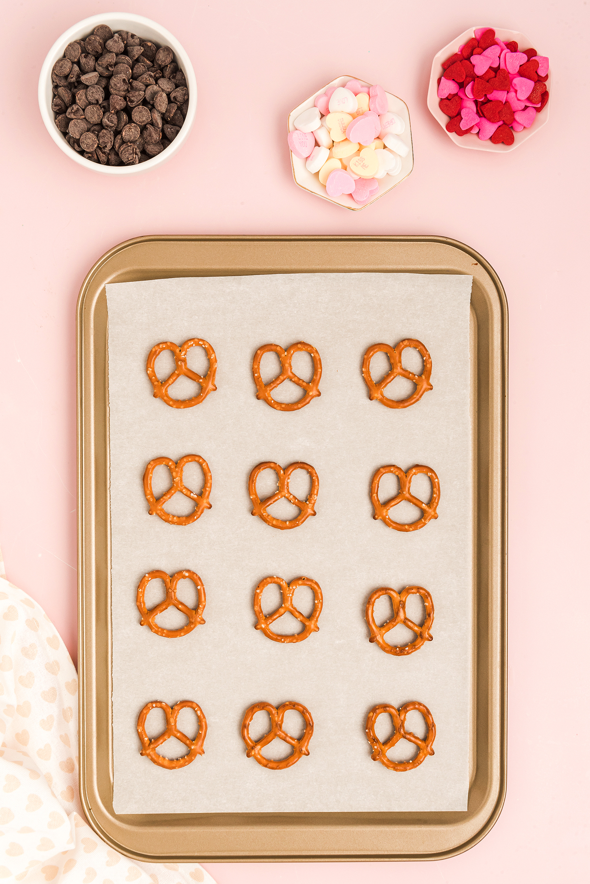 A baking sheet with Valentine Pretzel Snack Bites, surrounded by bowls of chocolate chips and heart-shaped candies on a pink background.