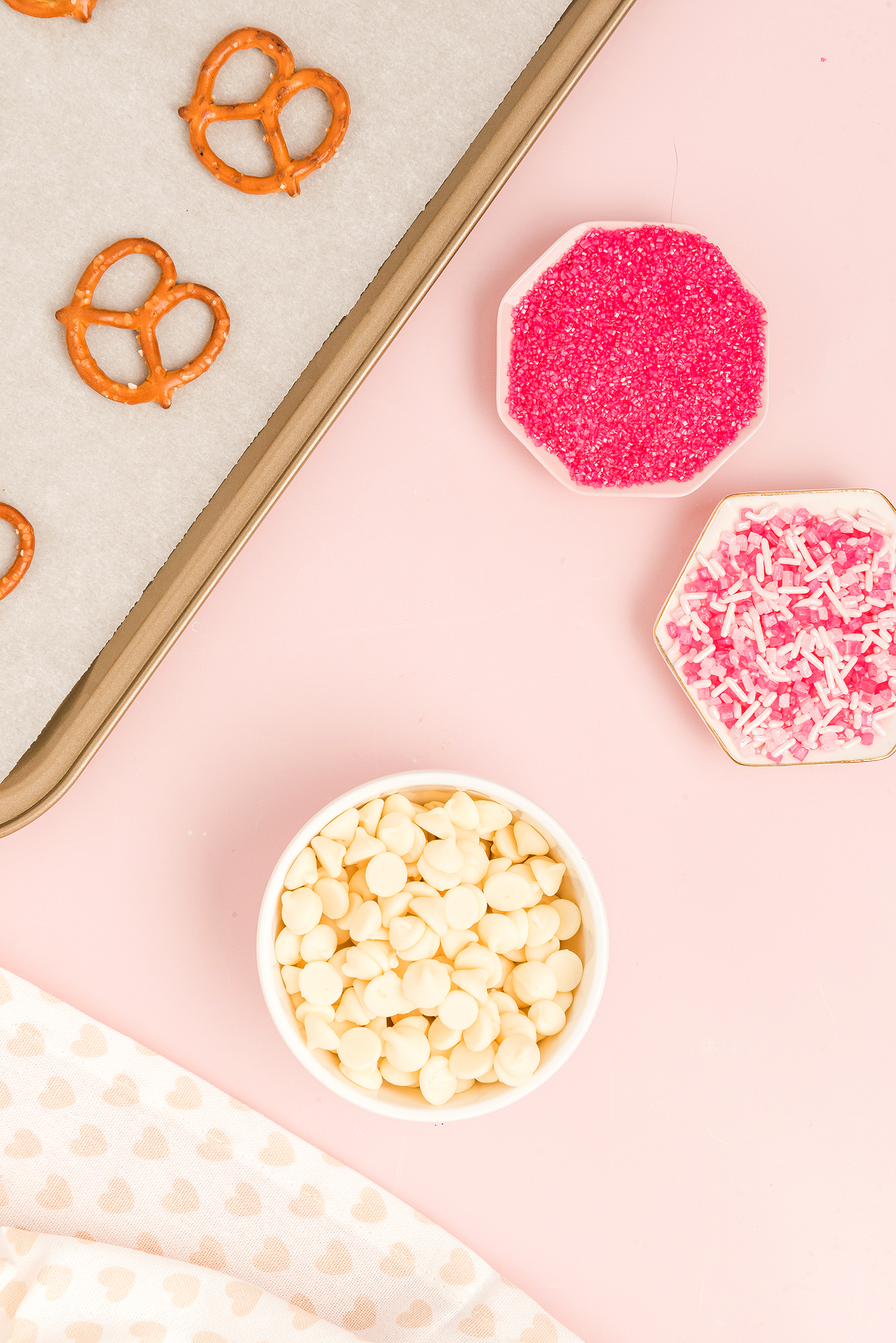 Baking tray with Valentine Pretzel Snack Bites, a bowl of white chocolate chips, and dishes of pink sprinkles and shredded coconut—perfect for a festive Valentine’s Day treat.