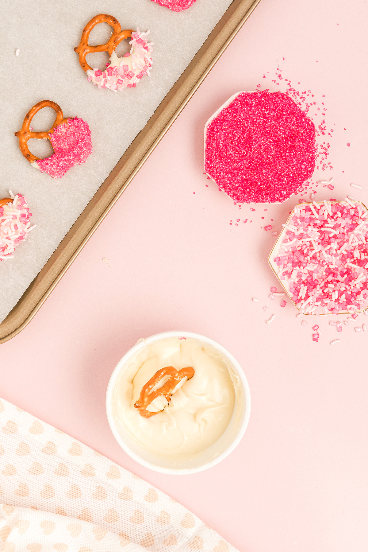 Pretzels with pink sprinkles and white dip on a pink surface make delightful Valentine Pretzel Snack Bites, presented with a baking tray and small dishes.