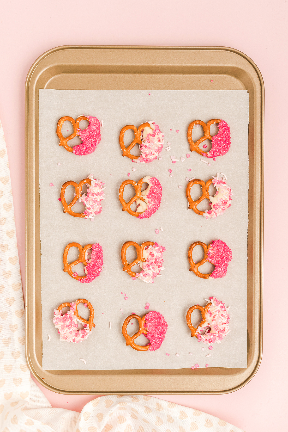 Baking sheet with Valentine pretzels dipped in white chocolate and pink sprinkles on parchment paper, set against a pink background—a sweet Valentine’s Day treat.