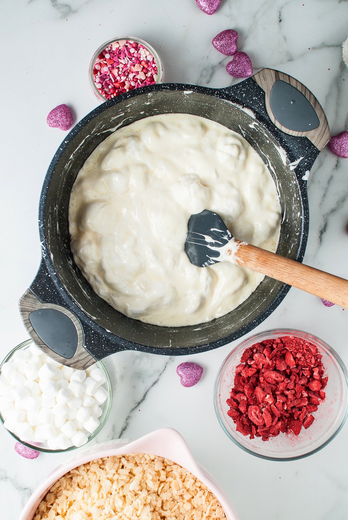 A pot of white melted mixture is stirred with a spatula, surrounded by bowls of sprinkles, marshmallows, berries, and freeze dried strawberries—perfect for making heart shaped rice krispie treats.