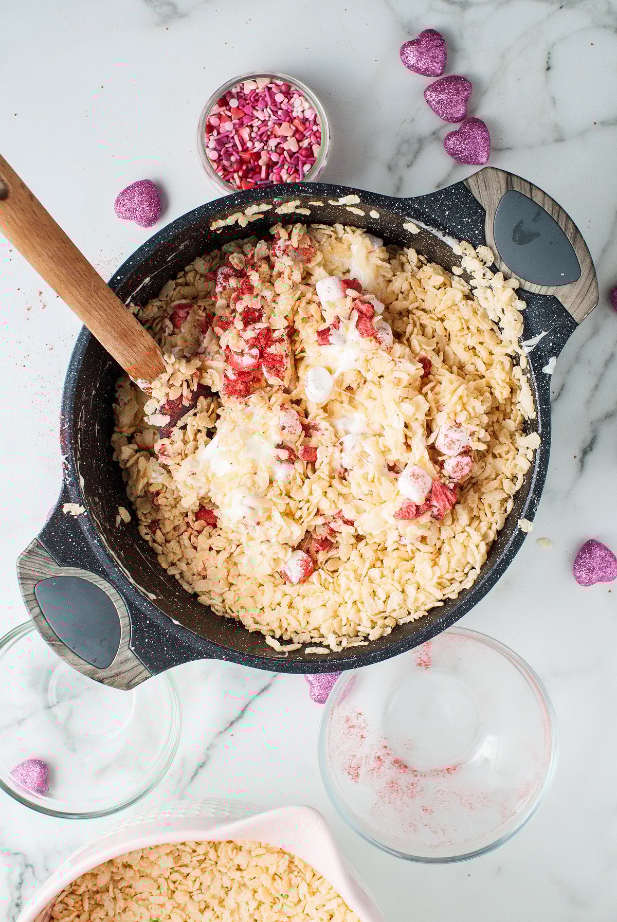 A pot of strawberry cream rice krispies mix with marshmallows and red sprinkles, surrounded by bowls and heart-shaped candies, creates a festive display of flavoured rice krispie treats.