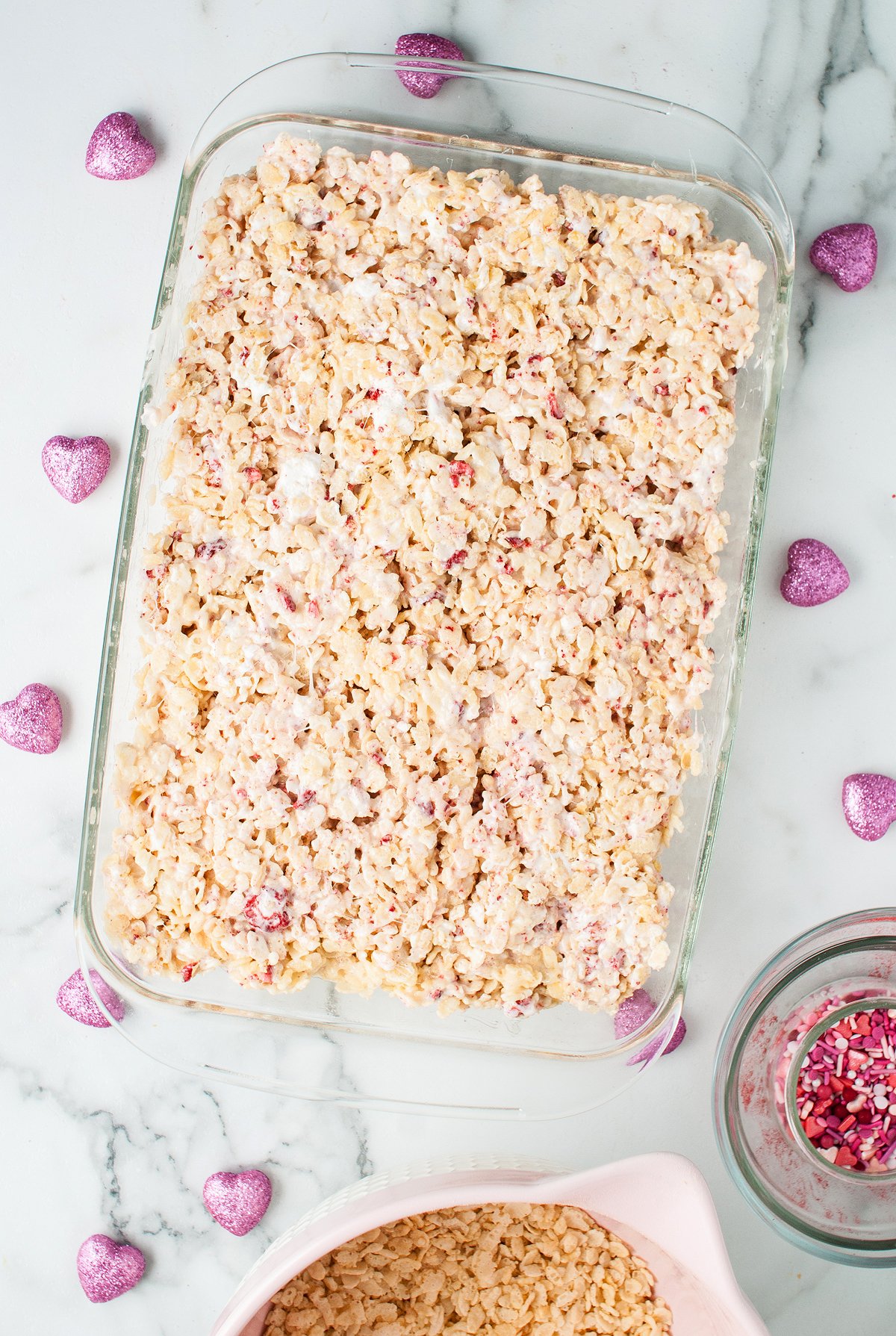 A glass dish of strawberry rice krispie treats on a marble surface, surrounded by pink heart-shaped decorations.