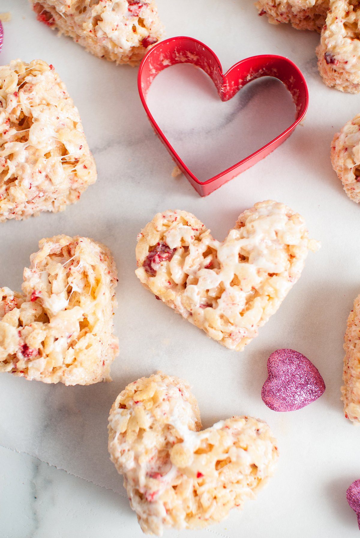 Heart-shaped rice krispie treats made with Strawberry Cream Rice Krispies, accented by freeze dried strawberries, a red heart-shaped cookie cutter, and pink glitter hearts on a white surface.
