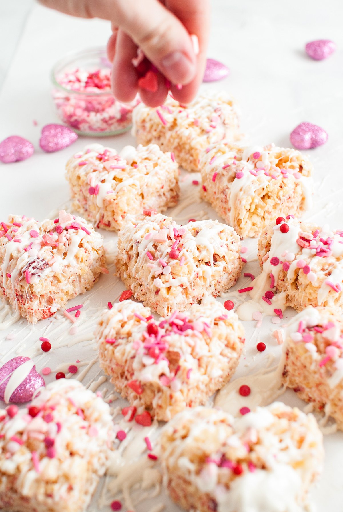 Heart-shaped rice krispie treats with white drizzle feature pink sprinkles and bits of freeze dried strawberries, added by hand for a sweet finishing touch.