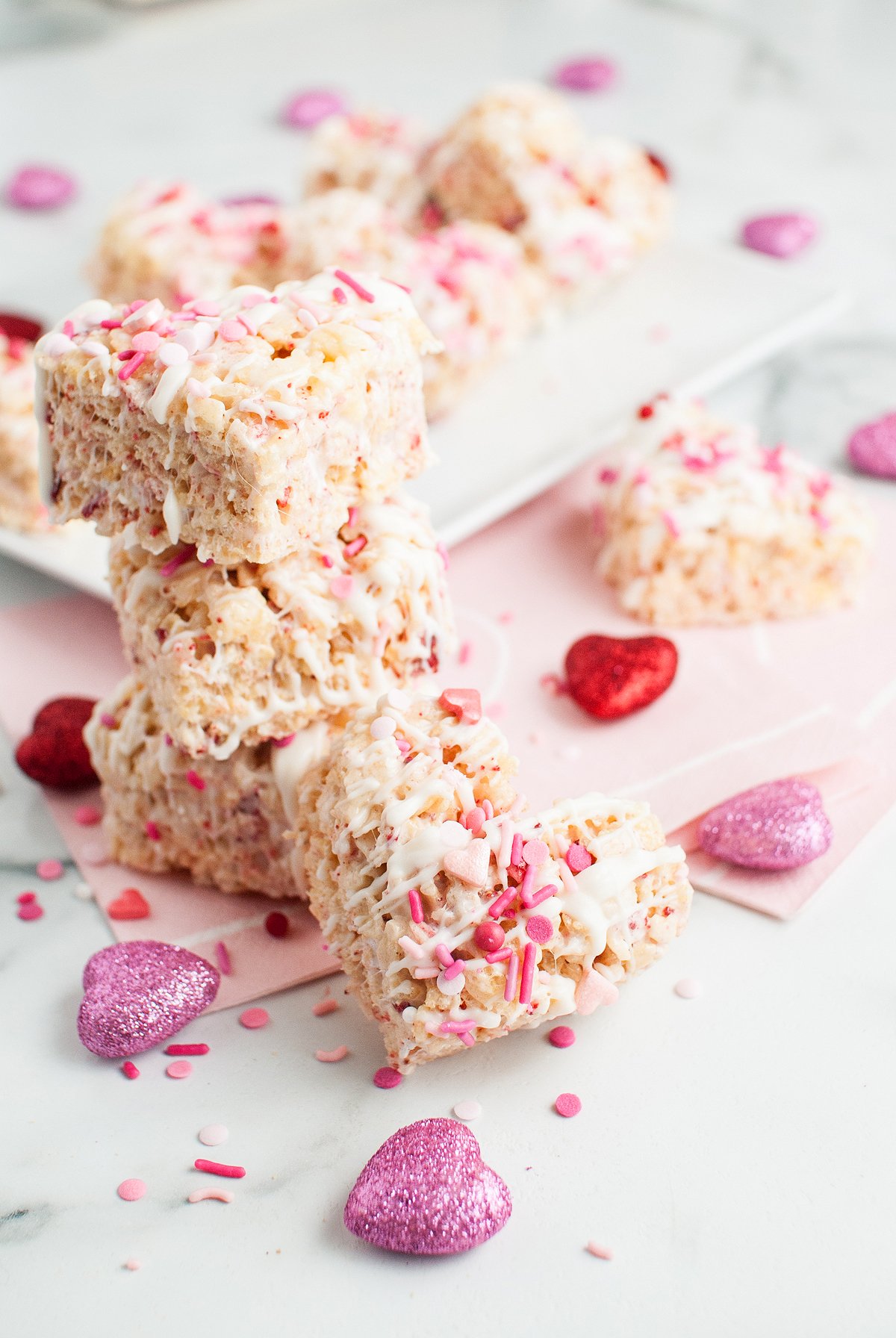 Strawberry rice krispie treats with pink sprinkles stacked on pink napkins, surrounded by glittery heart decorations and bits of freeze dried strawberries.