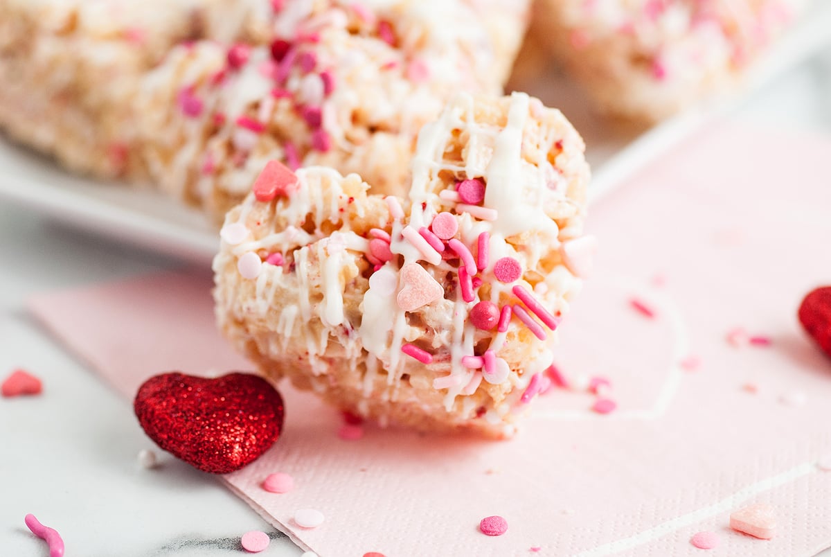 Heart-shaped Strawberry Cream Rice Krispies treat with pink, white, and heart sprinkles, drizzled with white icing and topped with bits of freeze dried strawberries.