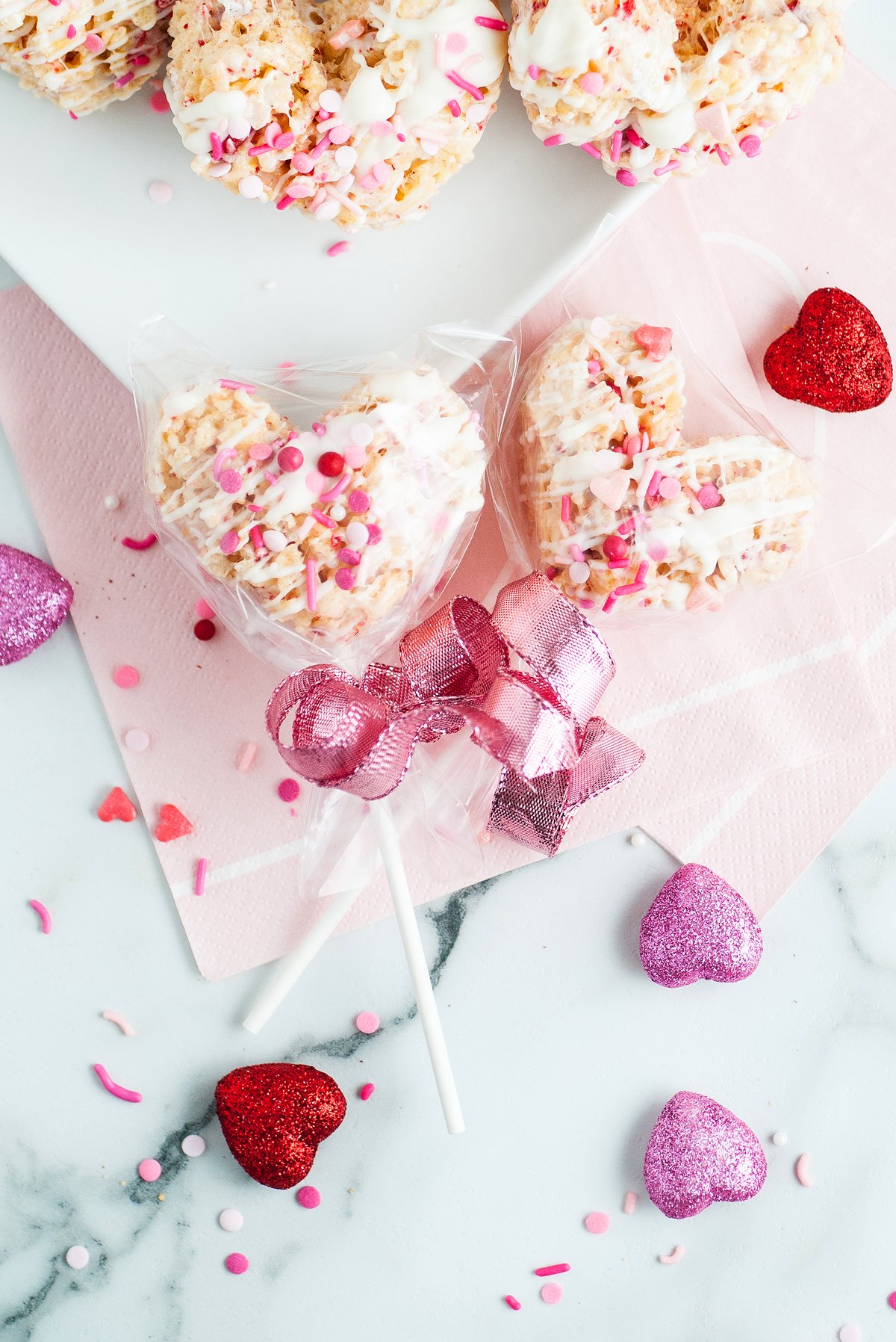 Heart-shaped strawberry rice krispie treats with pink sprinkles and ribbon, surrounded by glittery heart decorations. Made with freeze dried strawberries for a burst of fruity flavor.