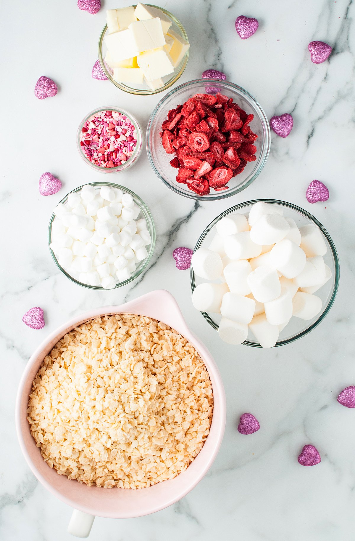 Bowls of rice cereal, marshmallows, butter, and freeze dried strawberries are ready to make delicious strawberry rice krispie treats, topped with pink sprinkles on a marble surface.