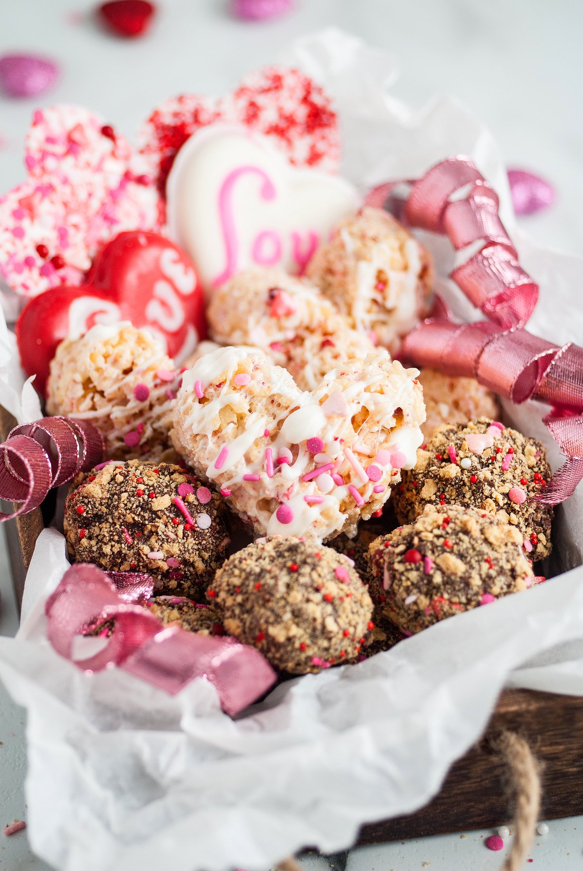 Assorted heart-shaped and round Valentine’s treats with pink ribbons, nestled in a wooden box lined with white paper—a charming Valentine’s sweets gift box made from easy recipes for a heartfelt surprise.