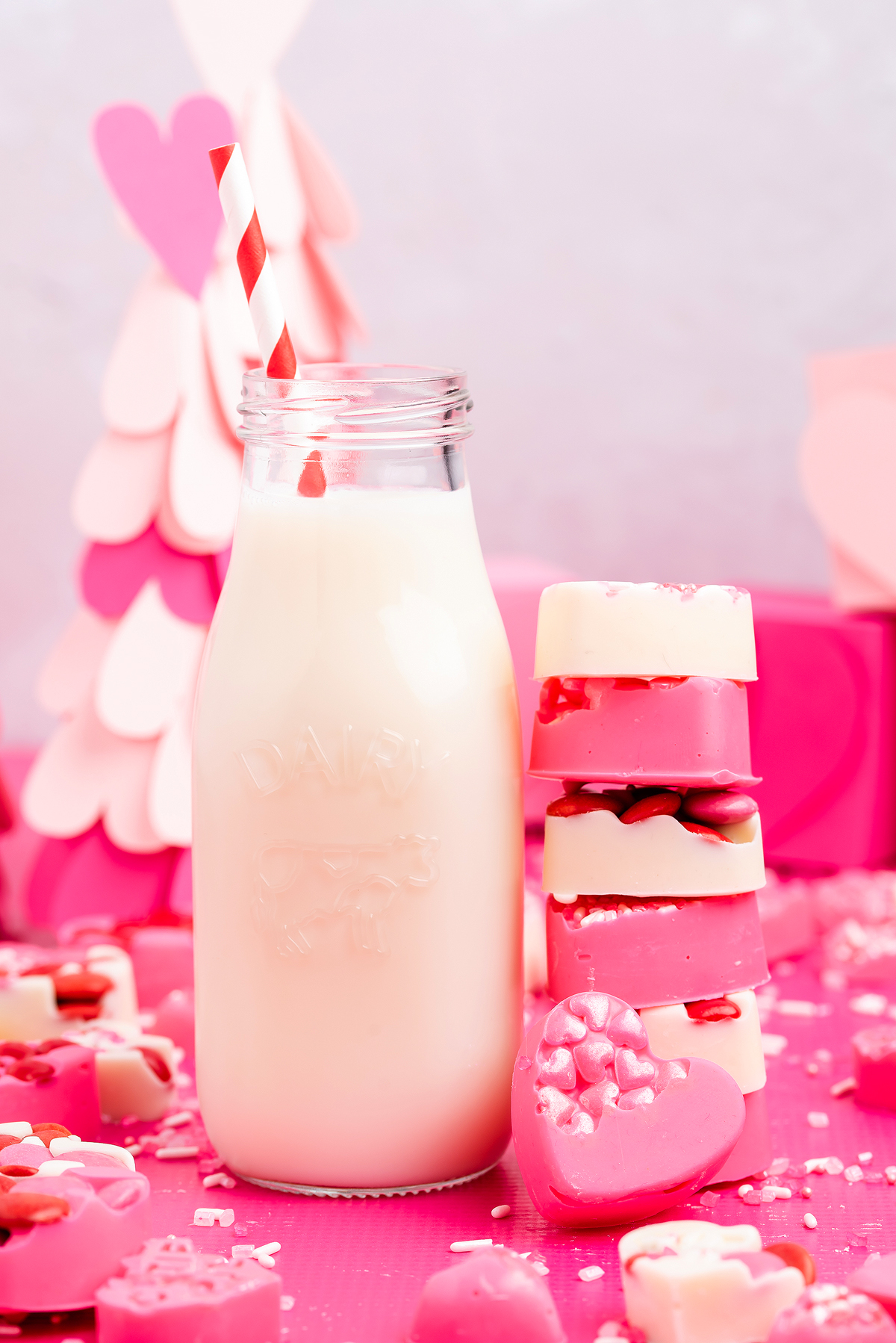 A glass bottle of milk with a red-striped straw, surrounded by pink and white heart-shaped candies and heart shaped chocolate treats—perfect for a DIY valentine chocolate gift.