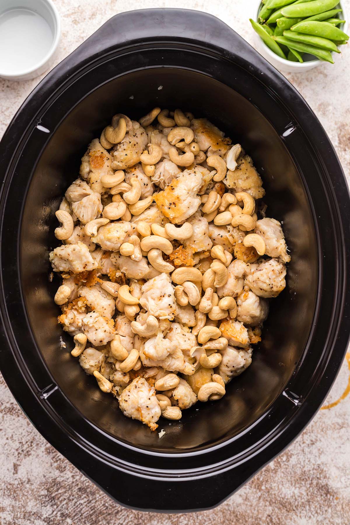 Chunks of chicken and cashews in a black slow cooker; snap peas in a bowl nearby.