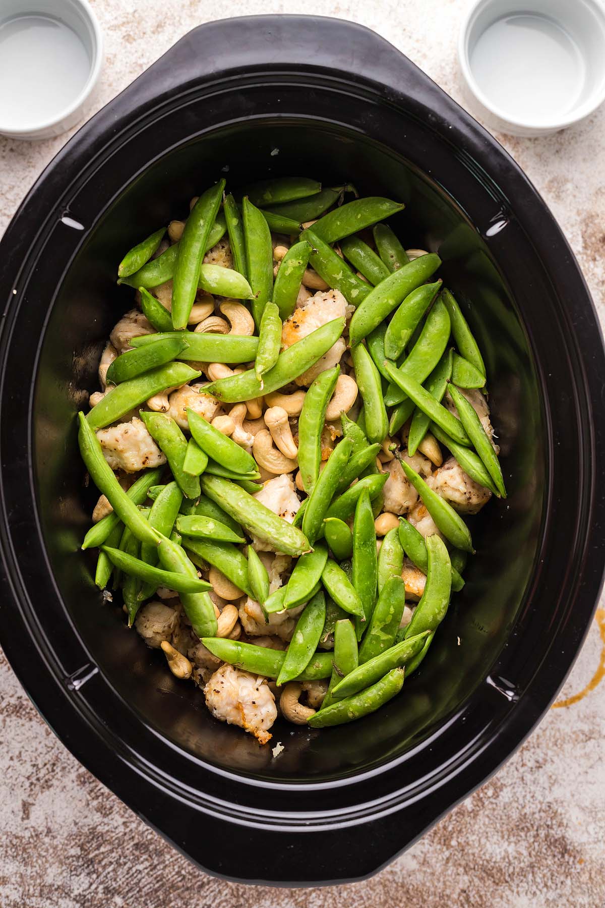 Chicken, snap peas, and cashews in a slow cooker, ready to be cooked. Two small bowls nearby.