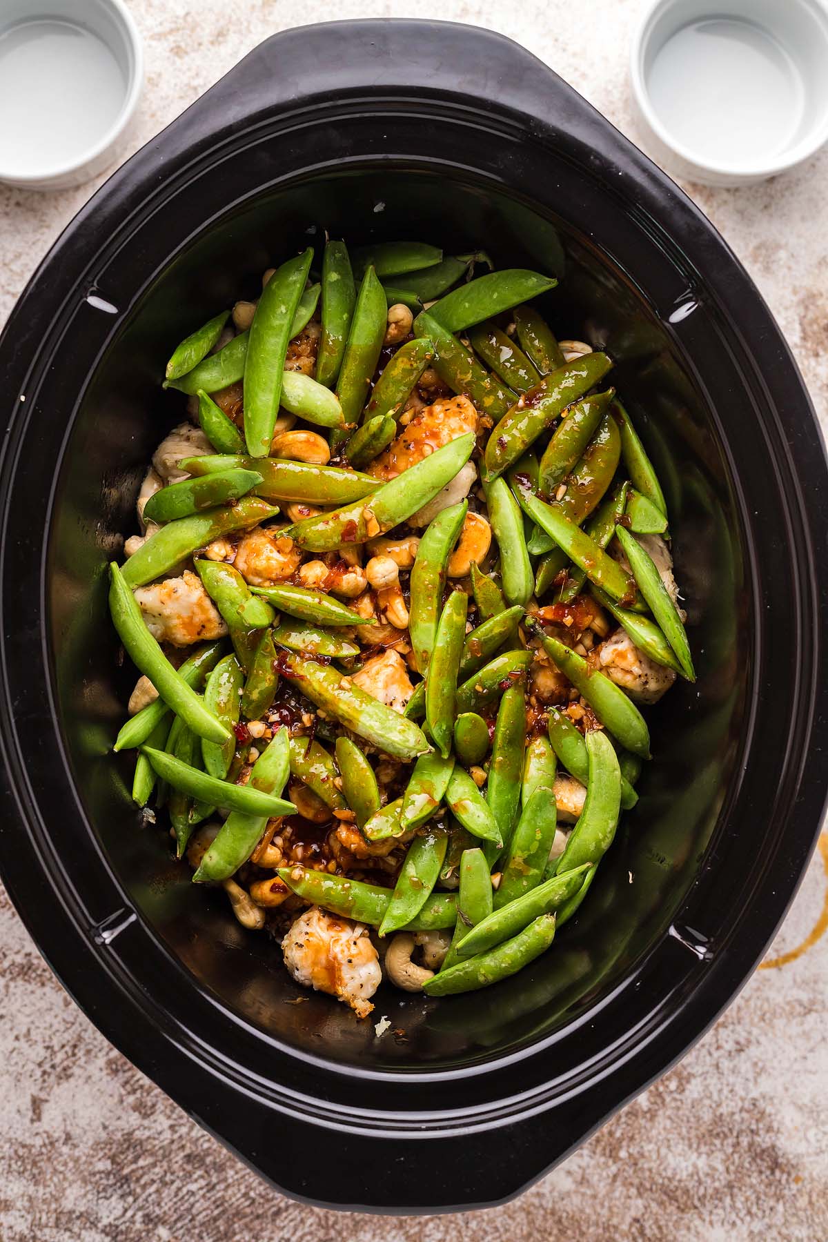 Chicken, snap peas, and cashews in a slow cooker, ready to be cooked. Two empty bowls beside the cooker.