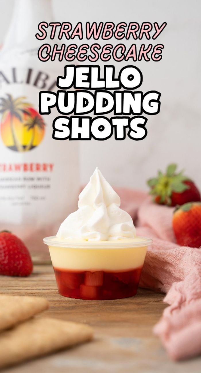 A Strawberry Cheesecake Jello Pudding Shot made with strawberry flavoured rum sits on a wooden table, topped with whipped cream and fresh strawberries.