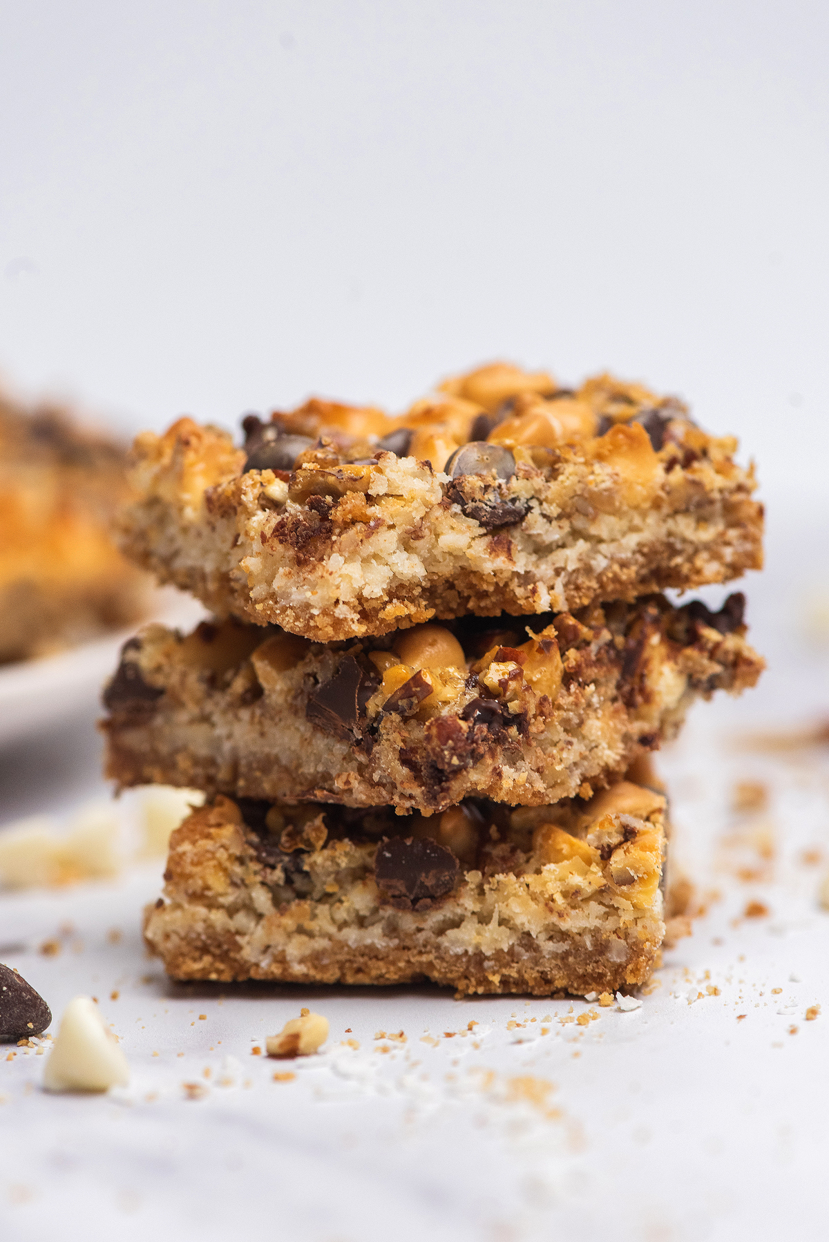 Three stacked chocolate coconut magic bars with butterscotch chips rest on a white surface, with crumbs scattered around.