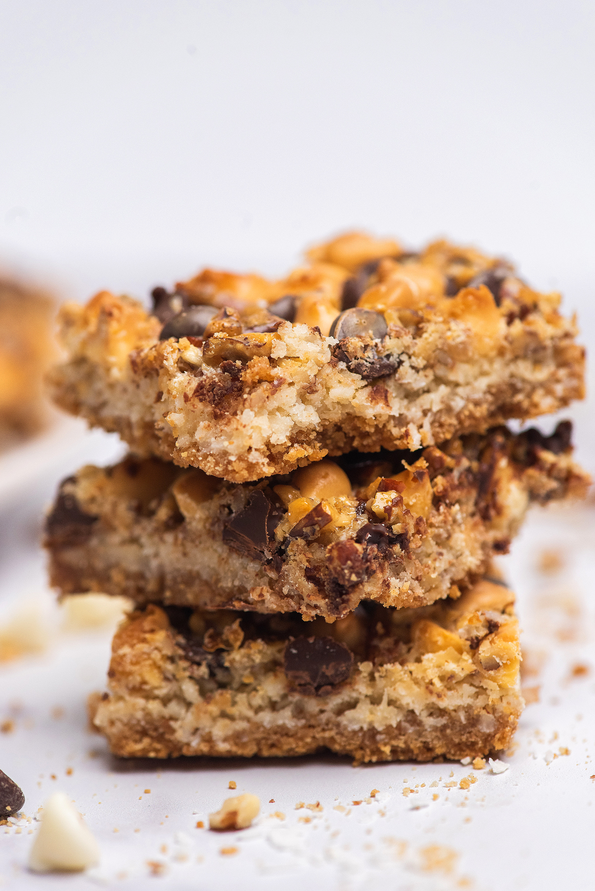 Three stacked Hello Dolly bars with chocolate chips and golden toppings rest on a white surface, surrounded by sweet crumbs.