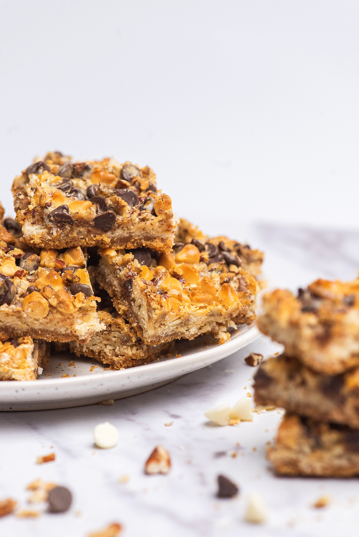 A plate of stacked Hello Dolly bars, also known as old fashioned coconut bars, topped with chocolate and butterscotch chips on a white surface.