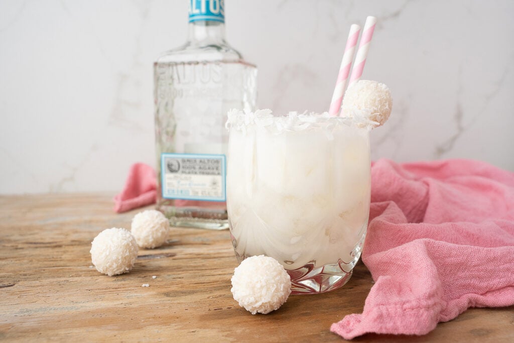 A coconut cocktail with straws and garnished with coconut truffles, next to a bottle of alcohol on a wooden surface with a pink cloth.
