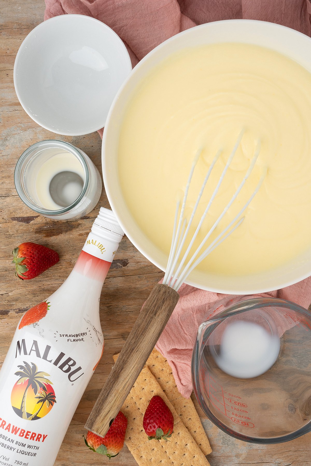 A bowl of creamy mixture with a whisk, Malibu bottle, strawberries, graham crackers, and measuring cups—perfect for making Strawberry Cheesecake Jello Pudding Shots.