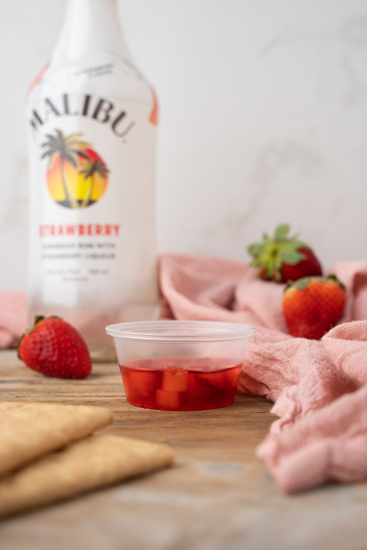A small cup of red gelatin with fruit, strawberries, and a Malibu bottle on a wooden table with a pink cloth—perfect for serving as strawberry pudding shots or a delightful strawberry dessert cocktail.