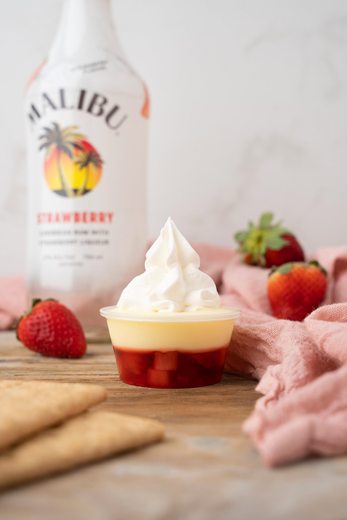 A dessert cup with layered strawberry, custard, and whipped cream, served as a playful strawberry pudding shot, with strawberries and a Malibu bottle behind.