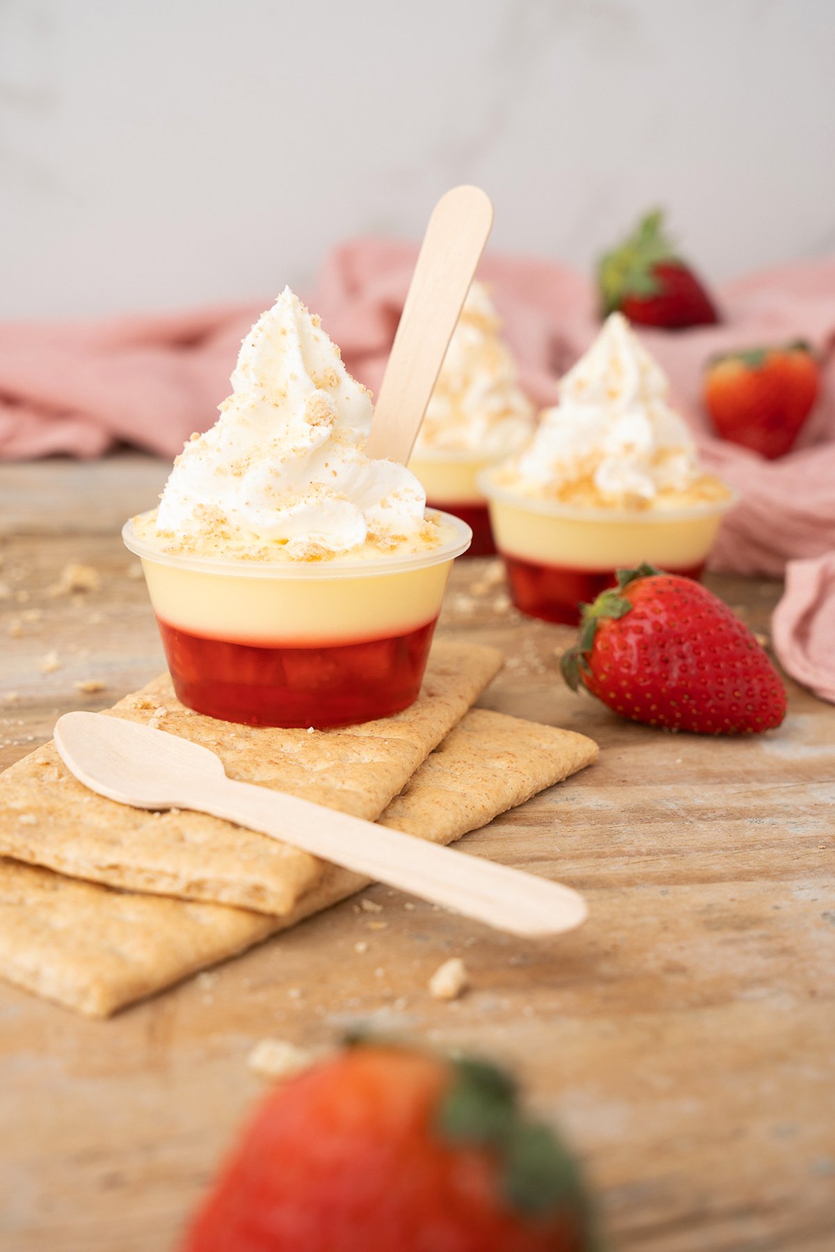 Mini strawberry parfaits with whipped cream on a wooden table, garnished with strawberries and graham crackers—perfect as a strawberry dessert cocktail inspired by Strawberry Cheesecake Jello Pudding Shots.